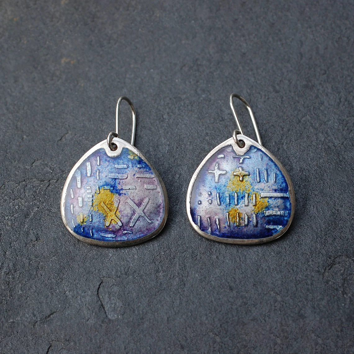 Fat teardrop shaped enamel earrings, etched silver, shades of purple and yellow. On a white grey slate background.