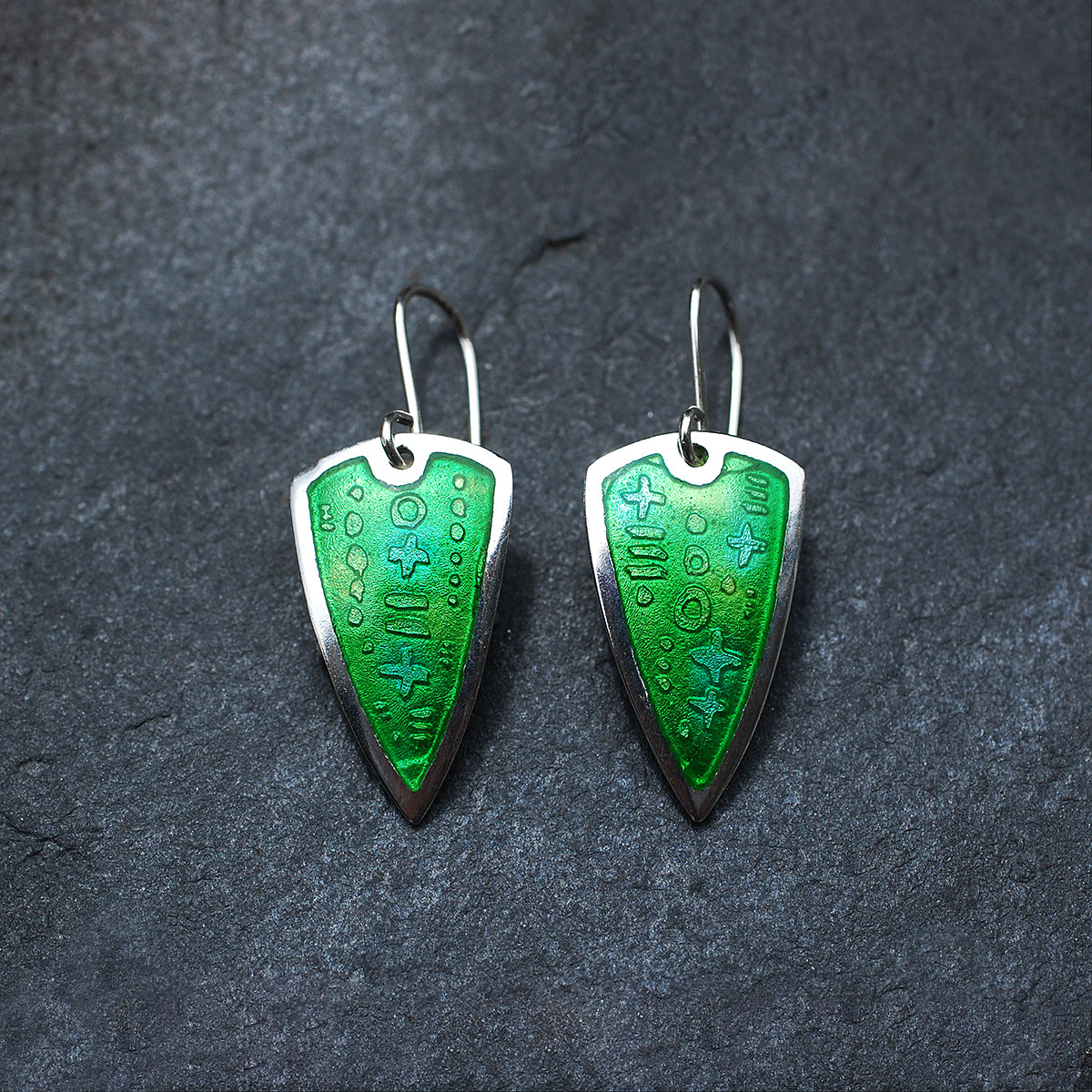 Silver earrings with green enamel over an etched pattern on a slate grey background.