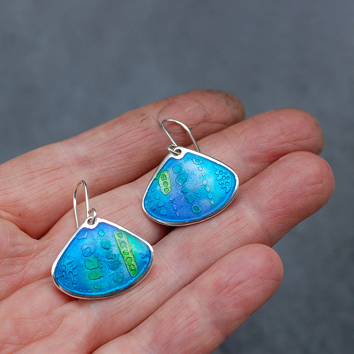 Onion shaped etched silver and enamel earrings in blues and greens, on a hand